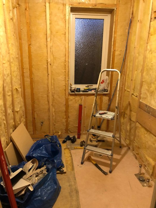Ett pågående badrumsrenoveringsprojekt med oskyddade väggar, isoleringsmaterial, en stege och byggmaterial utspridda på golvet.