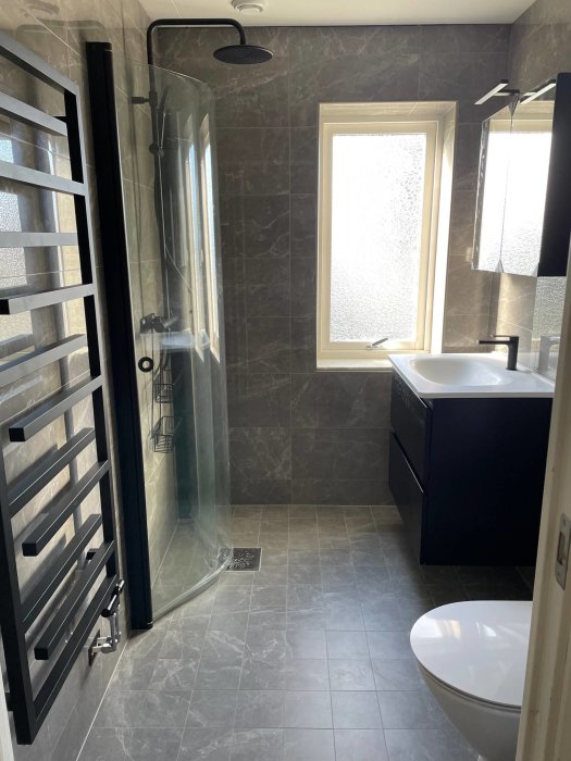 Renoverat badrum med gråa plattor, infälld dusch framför fönster, handdukstork och svart badrumsmöbel.