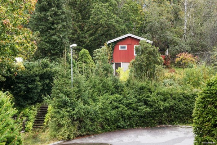 Vy från gatan av en röd stuga uppe på en kulle omgiven av grönska och en trappstegsledande uppgång.