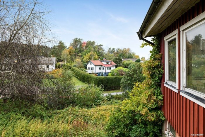 Vy från befintligt hus över en lutande tomt med ett annat hus i bakgrunden omgivet av grönska.