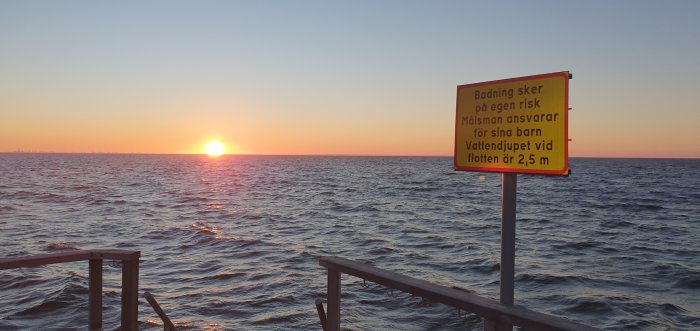 Skylt med badningsvarning vid havet i solnedgång, vågor och trätrapp.