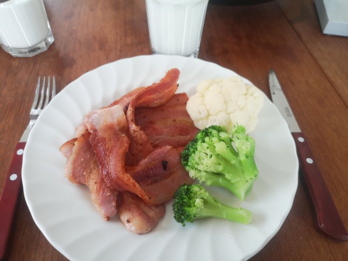 Delvis stekt bacon med blomkål och broccoli på en vit tallrik, med bestick och glas mjölk.