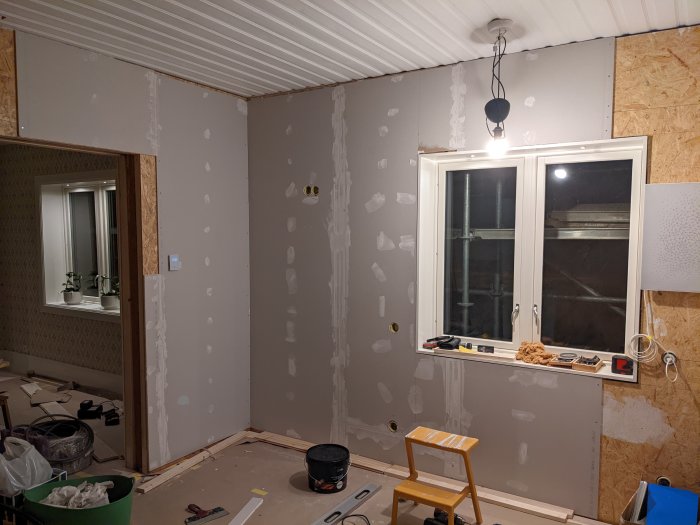 Rum under renovering med gipsskivor på väggar, spackling på skarvar och målade hyllor vid fönstret.