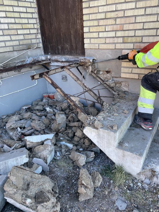 En person i arbetskläder river en betongtrappa med en elektrisk jackhammer, sten och murbruk på marken.