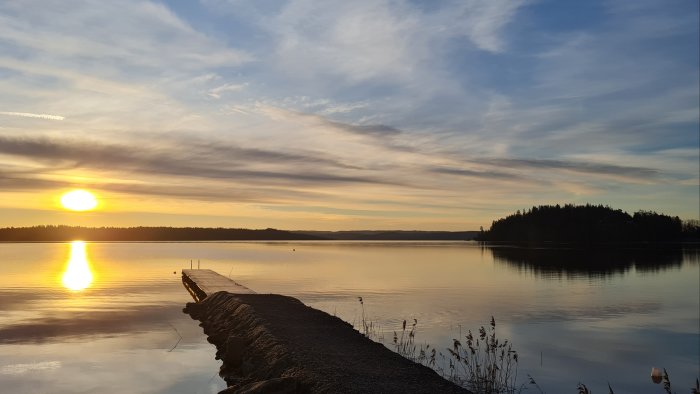 Soluppgång över en lugn sjö med brygga och träd i silhuett mot en himmel med moln.