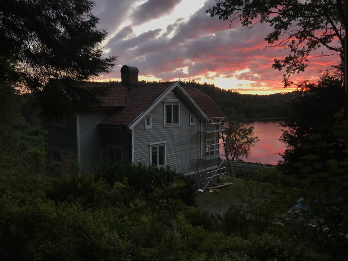 Renoverat hus vid sjö i skymningen med trädgård och solnedgång i bakgrunden.
