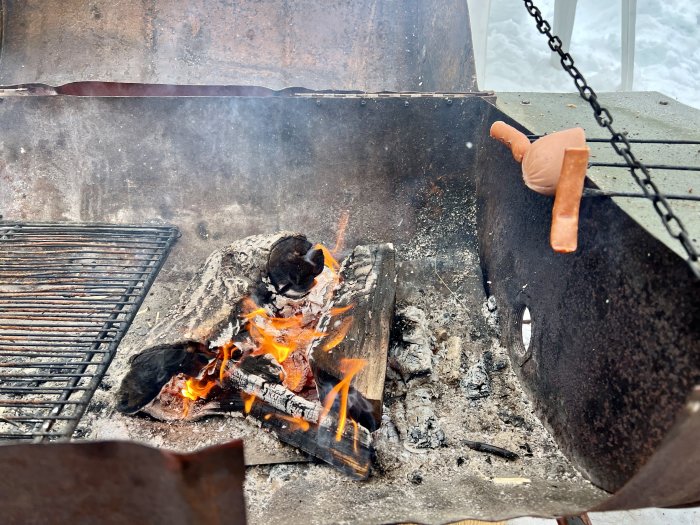 En korv lagas över en öppen eld i en rostig utomhusgrill med synliga lågor under vintern.