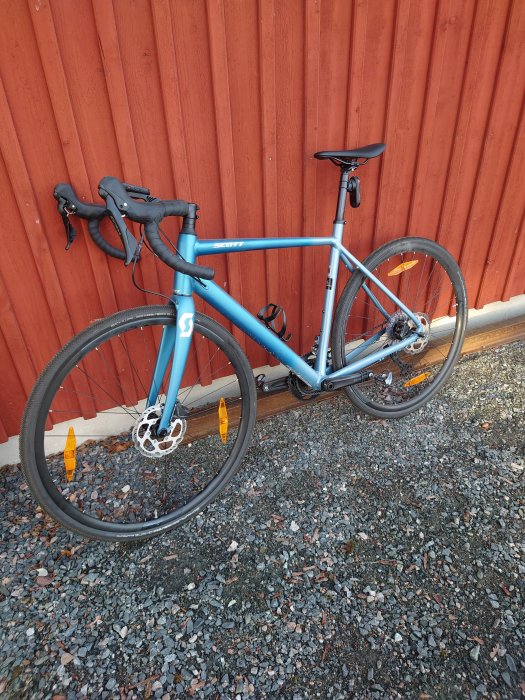 Blå landsvägscykel lutad mot röd trävägg på grusunderlag.