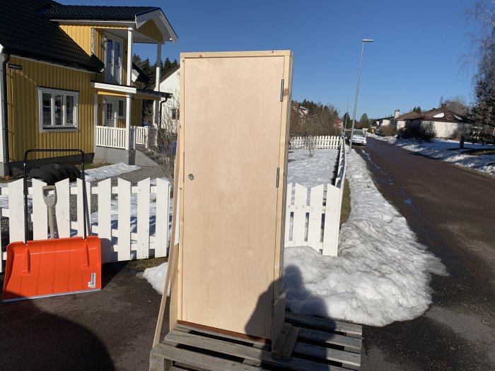 Nymålad dörr till matkällare står utomhus på en pall, med snö och bostadshus i bakgrunden.