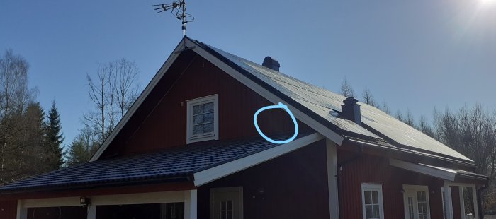 Rött hus med cirkelmarkerad ventilationsutgång på taket mot en klarblå himmel.