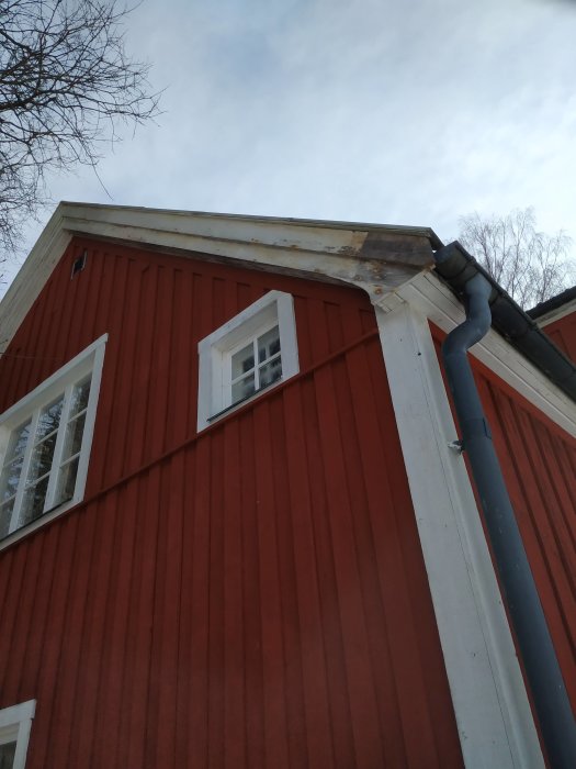 Hörn av rött trähus med sliten färg på vindskiva och husknut, fönster och stuprör syns.