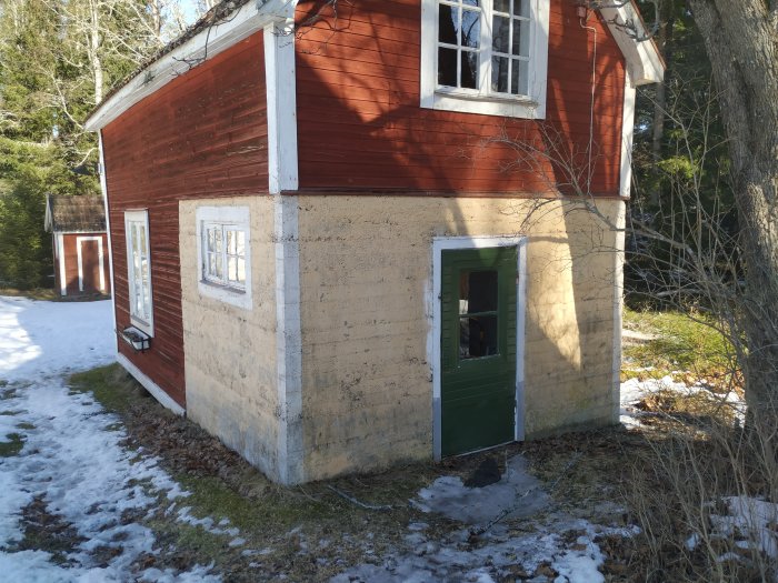 Gammalt rött torp med en putsad del i beige, omgivet av snötäckt mark och trädstammar.