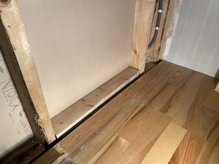 Hörn av ett rum där en mellanvägg möter trägolv, med synliga tecken på misstänkt svartmögel vid basen av väggen.