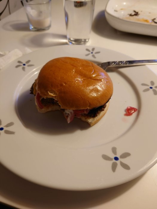 Halväten hamburgare med ketchup på ett vitt blommönstrat fat vid sidan av tomma tallrikar och glas.