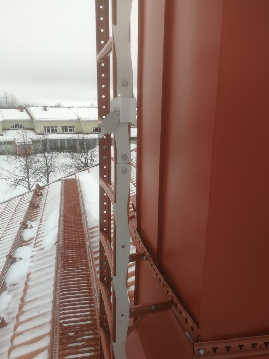 Omålad stålställning fäst vid röda stolpar för fallskydd på hus med snötäckt tak i bakgrunden.