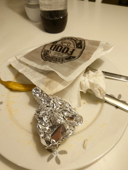 Resterna av en måltid från Romme pizzeria med en folieinpackad bit, servetter och en flaska på ett smutsigt vitt fat.