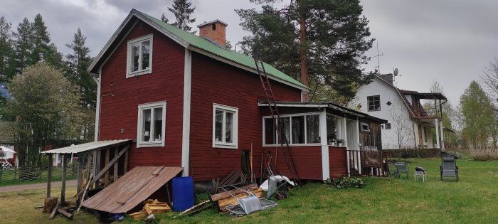 Rött hus med vit knut, tillbyggt uterum i vitt och en carport, omgivet av trädgårdsröra och verktyg.