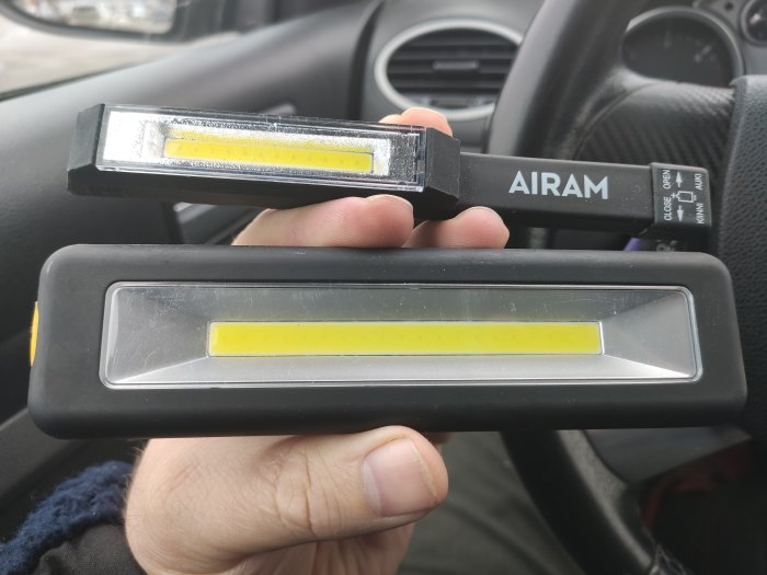 Hand håller två bärbara arbetslampor med LED-ljuskällor i ett bilens förarsäte.