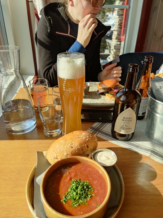 Lunchbord med gulasch i skål, bröd, gräddfil och en ölsejdel, bakgrunden visar en person och en hamburgermål.