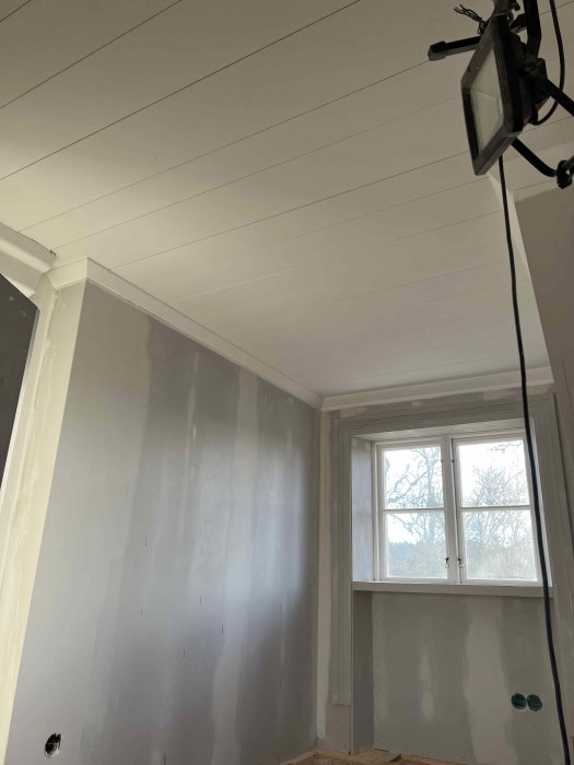 Nymålat vit tak i ett renoveringsrum med oslipade väggar och ett fönster.