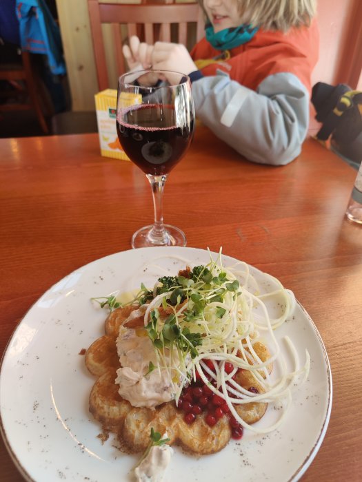 Vällagad maträtt på tallrik med ett glas rödvin och någon som skålar i bakgrunden.