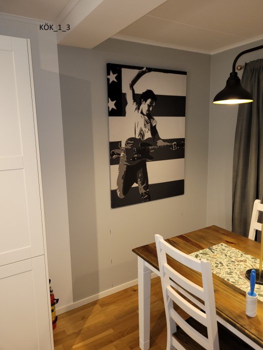 Hörn av ett kök med vit och ljusgrå inredning, matsalsbord och stol, samt en stor svartvit tavla på väggen.
