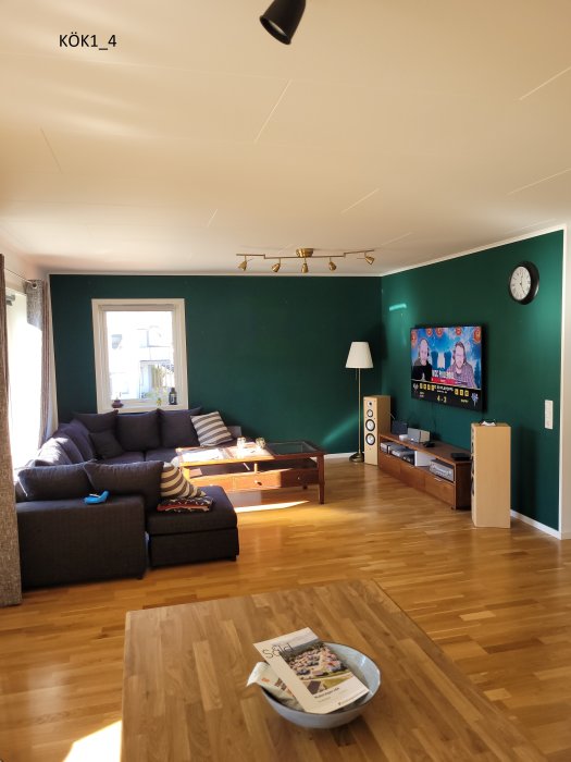 Vardagsrum med grön vägg, grå soffa och TV, trägolv, vit tak och belysning.
