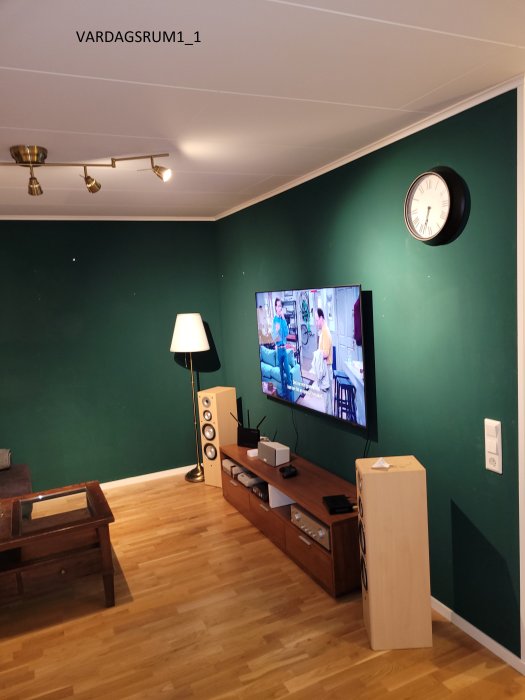 Vardagsrum med mörkgrön vägg, vit tak, brunt golv, TV på väggen och brun möblering.