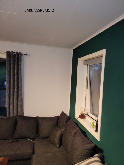 Vardagsrum med grön vägg, grå soffa och vitt fönster med gardiner.