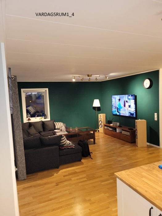 Vardagsrum med gröna väggar, tv, soffa och trägolv synligt från ett kök.