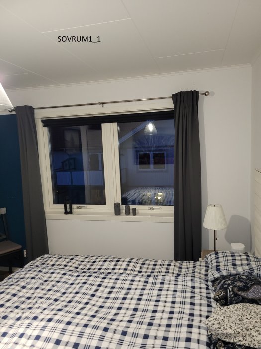 Ett sovrum med vit väggar, mörkblå gardiner, rutigt överkast på sängen och fönster som visar utomhusvy.