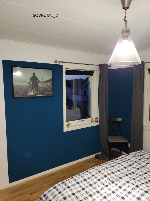 Sovrum med blåmålad vägg, vit tak, trägolv, grå gardiner, fönster och inramad bild.