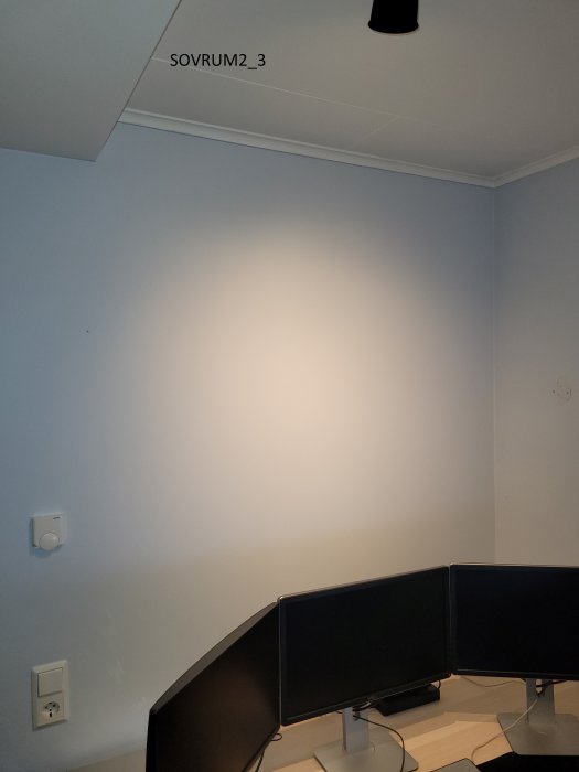 Hörn av ett rum med babyblå väggar, datorskärmar på skrivbord och taklampa.