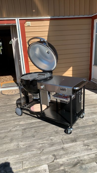 Weber Charcoal Summit-grill öppen och parkerad på en träaltan utanför en röd husvägg.