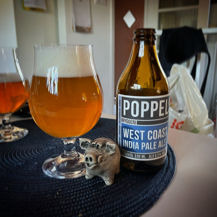 Ölglas och flaska av Poppels West Coast IPA på ett bord med en smågrisfigur.