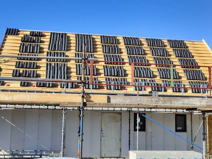 Delvis lagt tak med svarta takpannor och röda ventilationsuttag på en byggställning.