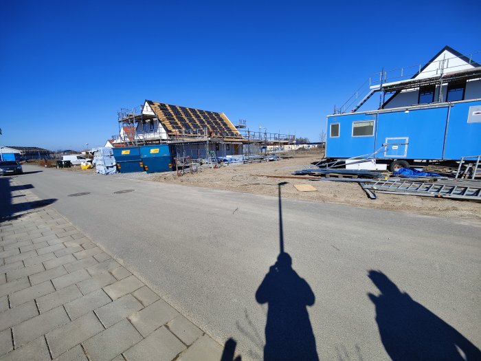 Hus under konstruktion med delvis lagt tak och rött ventilationsmunstycke, skugga på marken, byggbodar i förgrunden.