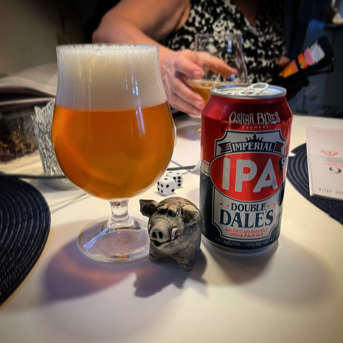 Ölglas fyllt med IPA bredvid en burk Double Dale's IPA, tärningar och en grishantverk på ett bord.