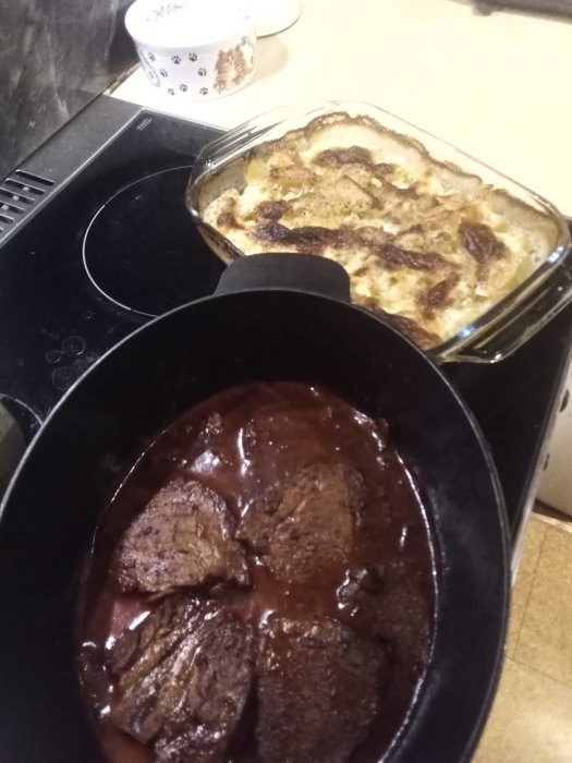 Gjord gratäng i en glasform och kött i brunsås i en gjutjärnspanna på spishäll.