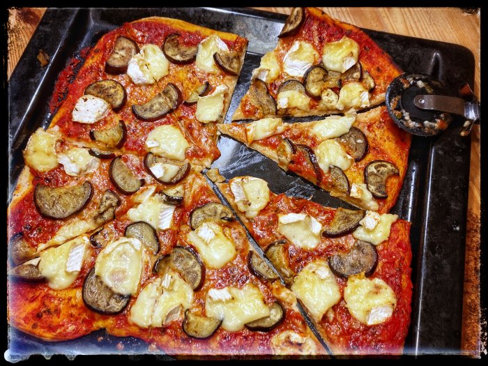 Hembakad pizza med tre ostar och aubergine på bakplåt, delvis uppäten, pizzaskärare i hörnet.