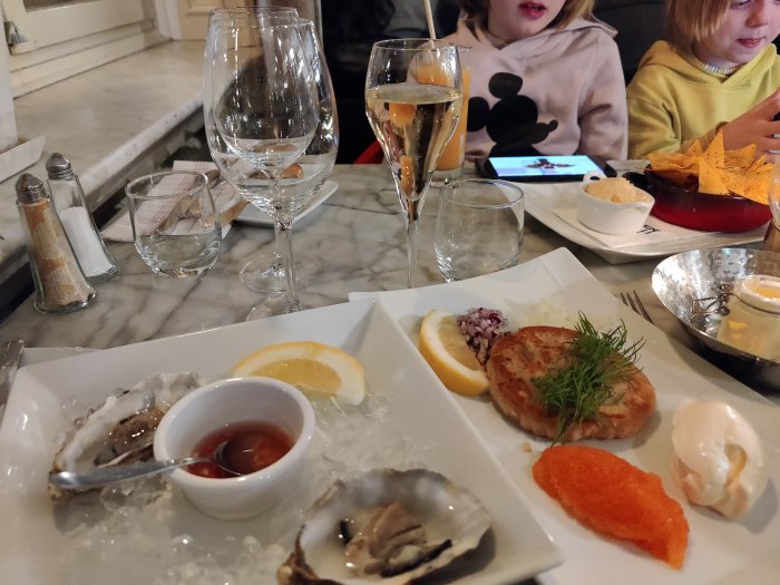 Förrättsfat med ostron och löjrom, citronskiva och tillbehör, med vinglas och personer i bakgrunden.