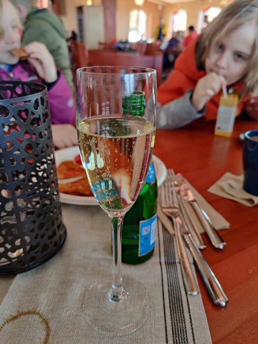 Ett glas mousserande vin framför barn som äter lunch inomhus.