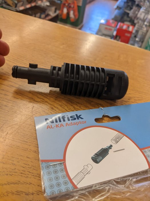 Svart Nilfisk AL-KA adapter i förgrunden med dess förpackning, på en butiksdisken.