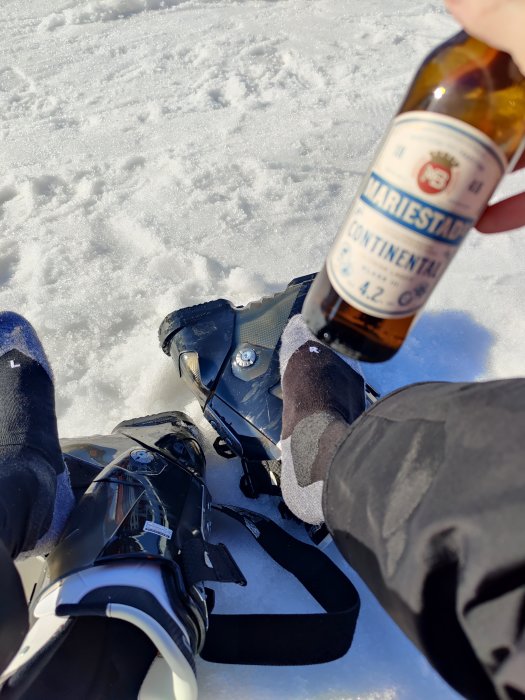 Person som håller en ölflaska med skidpjäxor och snö i bakgrunden.