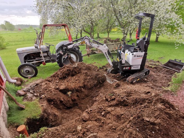 Grävningsarbete med en Bobcat minigrävare bredvid en äldre traktor, grävd grop och avloppsrör i trädgård.