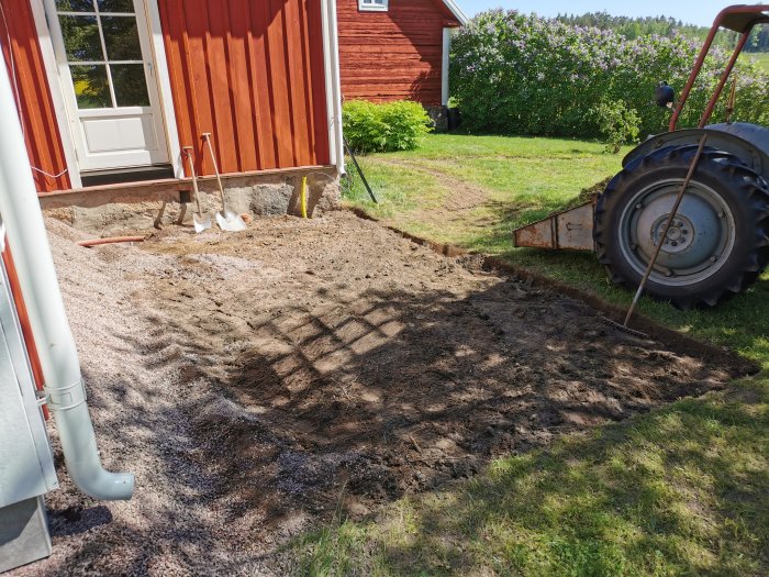 Förberedelse av mark med grus och markduk för installation av bad, bredvid röd stuga och grönt gräs.