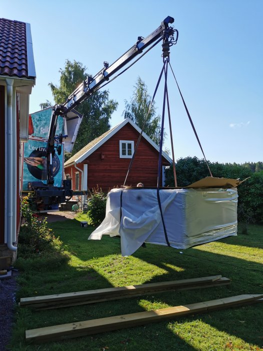 Badkar omslutet av presenning lyfts av kran i trädgård med träreglar på marken för altanbygge.