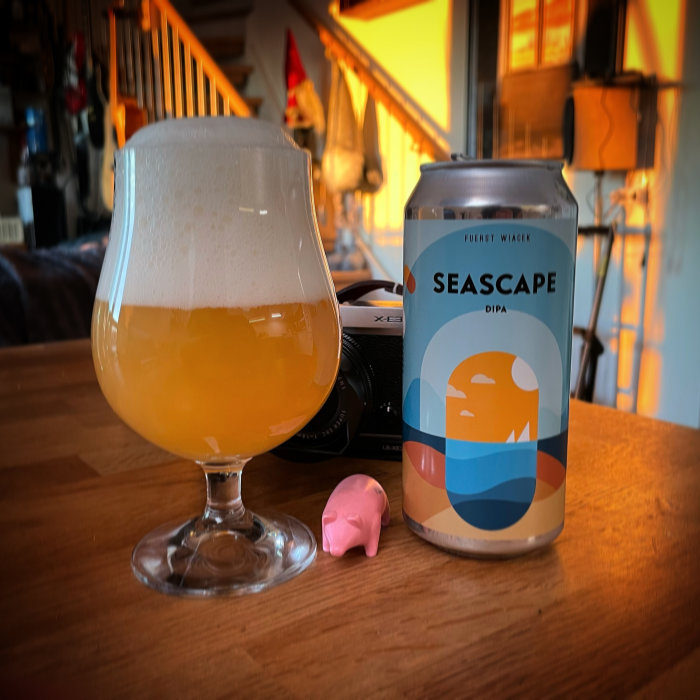 Ölglas fyllt med skummande öl bredvid en burk med etiketten "SEASCAPE DIPA", kamera och liten rosa figur på ett bord.