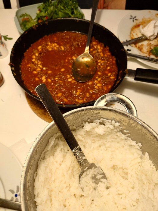 Kokad ris i en kastrull bredvid en gryta med chili con carne och serveringssked. Resterna av en måltid syns.