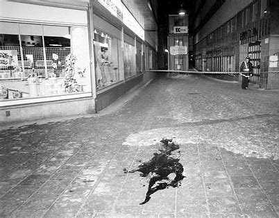 Svartvit bild från 1986 av en tom gata med affärer och en stor mörk fläck på marken.